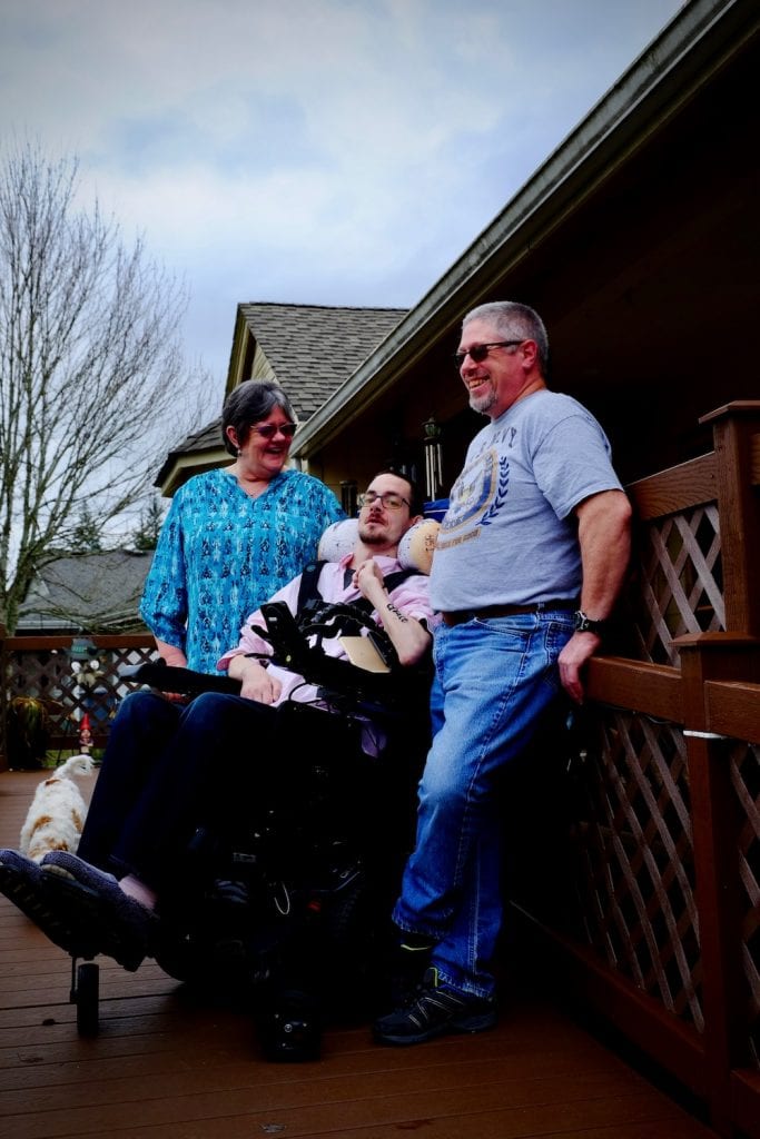 Photo of clients:Three individuals are on a ramp in front of their home. It's a cloudy day and there is a tree in background. The woman on the left is smiling and looking at her son in the middle who uses a wheelchair. His father on the right is smiling and looking towards the street.